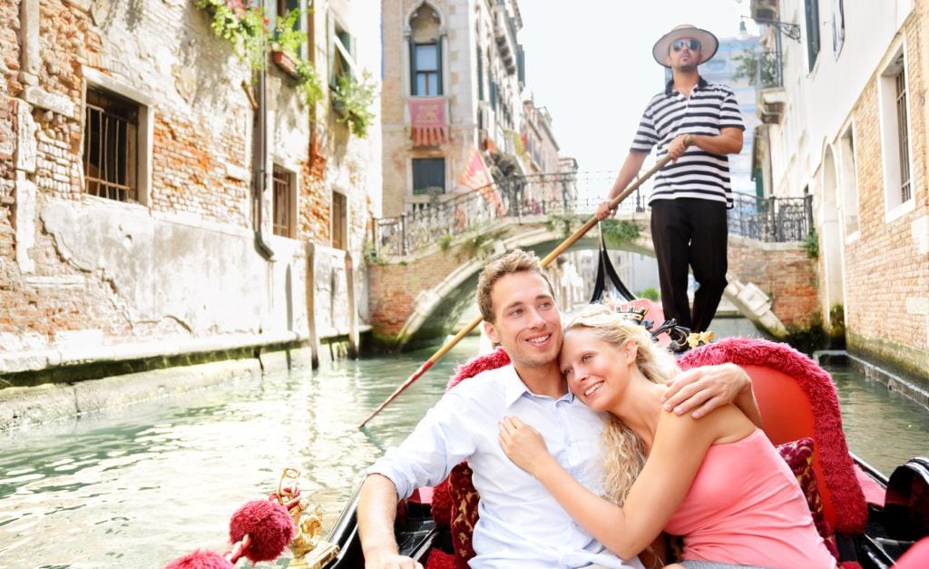 Romantic travel couple in Venice on Gondole ride romance in boat happy together on travel vacation holidays. Romantic young beautiful couple sailing in venetian canal in gondola. Italy, Europe.